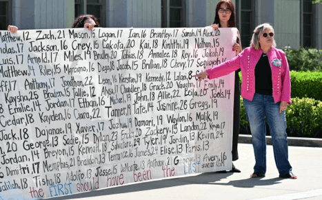 A woman in a pink jacket with a large button of a teenage girl affixed to it stands in front of a large banner with the names and ages of youth who have been victims of social media.
