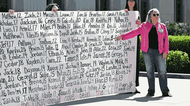 A woman in a pink jacket with a large button of a teenage girl affixed to it stands in front of a large banner with the names and ages of youth who have been victims of social media.